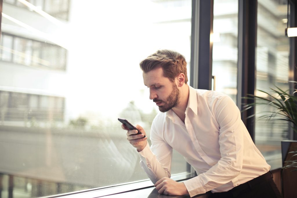 pexels-photo-859265-859265 A businessman in a white shirt texts on his smartphone by a large glass window inside an office.