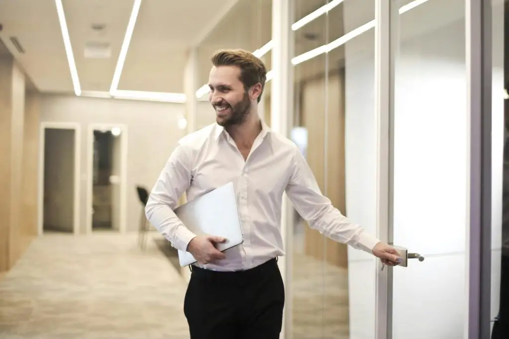 pexels-photo-901424-901424 A smiling man holds a laptop while standing in a modern office hallway, exuding confidence.